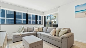 A modern living room with a large sectional sofa, a small ottoman, and a large window with multiple panes. - Allura Bella, Jervis Bay (Huskisson)