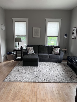 A living room with a dark sofa, a patterned rug, and a wooden floor. - The Grand Carriage Townhouse (Eau Claire)