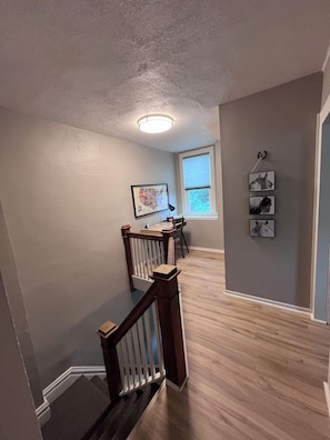 A staircase with wooden railings leading to a room with a desk and chair, a window with blinds, and wall-mounted artwork. - The Grand Carriage Townhouse (Eau Claire)