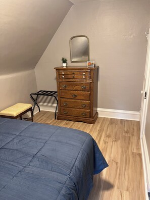 A bedroom with a wooden dresser, a bed with a blue bedspread, a small stool, and a mirror on the dresser. - The Grand Carriage Townhouse (Eau Claire)