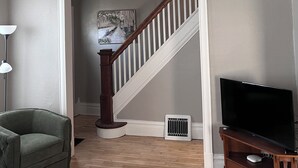 A living room with a staircase, a green armchair, a wooden entertainment center, and a flat-screen TV. - The Hudson Carriage Townhouse (Eau Claire)