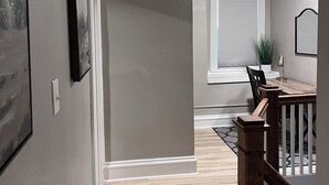 A hallway with a wooden floor, a dining area with a table and chairs, and a staircase with a railing. - The Hudson Carriage Townhouse (Eau Claire)