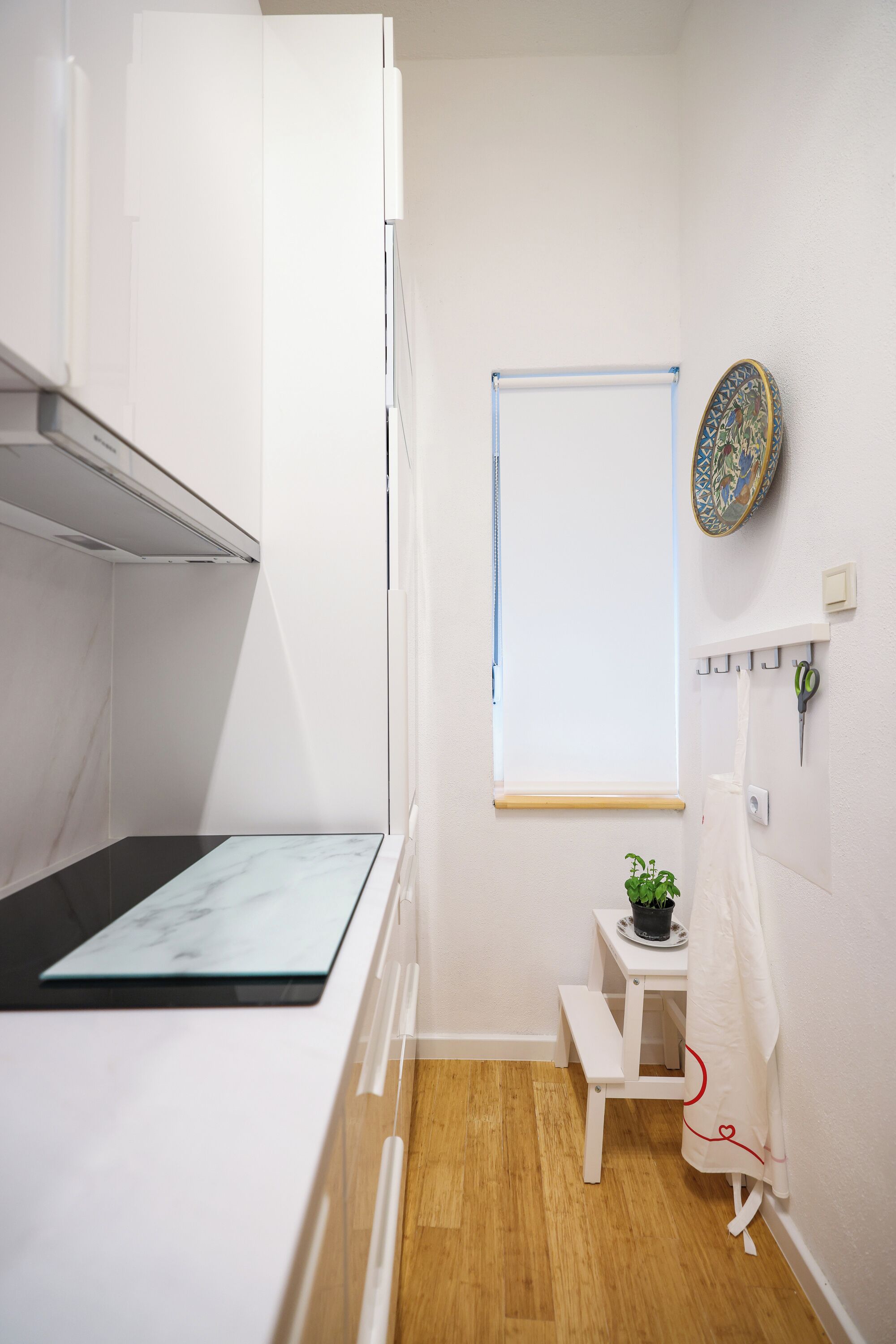 A compact, modern room with a kitchenette, a small table with a plant, a whiteboard, and a decorative plate on the wall.