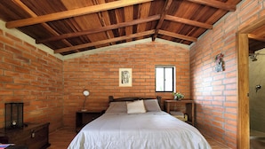 A bedroom with a brick wall, wooden ceiling beams, a bed with a white bedspread, a nightstand with a lamp, and a window. - Nutabe House, Girardota 40 min from Medellín! Ideal house for families and friends  (Girardota)