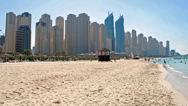 Ein sandiger Strand mit einem Rettungsschwimmturm und Sonnenschirmen, im Hintergrund ein Stadtbild mit hohen Gebäuden.