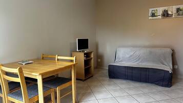 Una habitación pequeña con una mesa de comedor de madera, sillas, un televisor sobre un mueble bajo y un sofá tapizado.