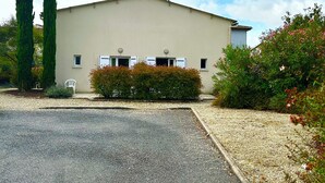 A single-story building with a flat roof, a gravel driveway, and a small garden area with shrubs and a chair. - Résidence Les Roussets (Jonzac)
