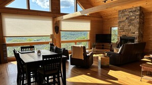 A wooden interior with a dining table, chairs, a fireplace, and large windows overlooking a forest. - Escape to a one-of-a-kind Mountaintop Retreat with panoramic westerly views (New Milford)