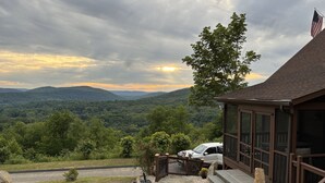 A cabin with a stone patio, a car parked in front, and a view of a forested mountain range. - Escape to a one-of-a-kind Mountaintop Retreat with panoramic westerly views (New Milford)