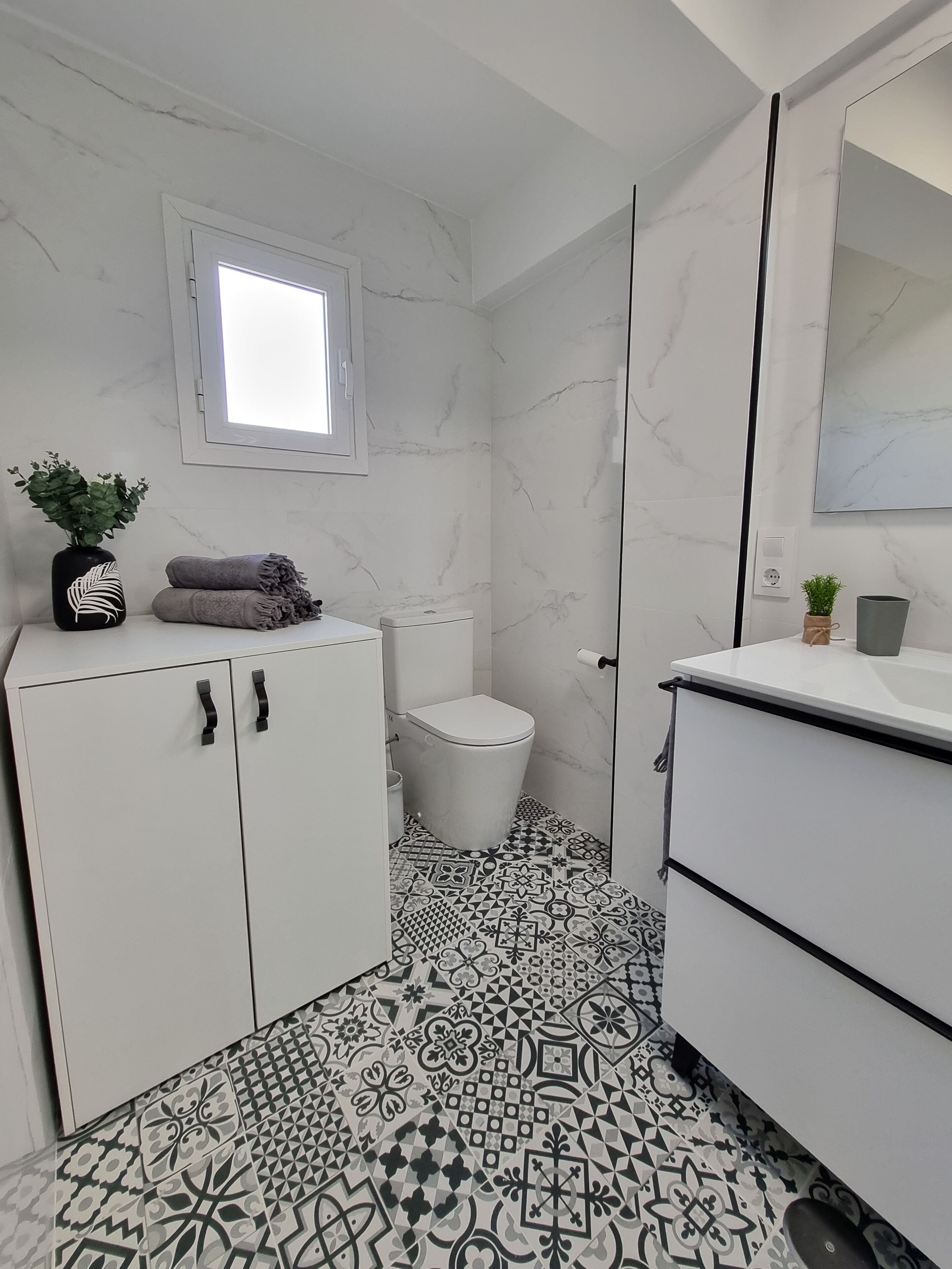 A bathroom with a white toilet, a sink, and a cabinet. There is a window above the toilet, and the floor has a patterned tile design.
