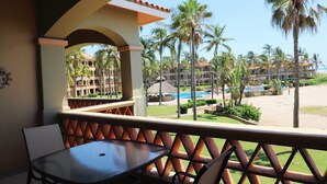 Un balcone con tavolo e sedie, vista su una piscina e palme, con un edificio di un resort sullo sfondo.