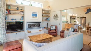 Un soggiorno moderno con un divano, poltrone, un tavolino da caffè e una televisione. Si vedono librerie piene di libri e oggetti decorativi. Sullo sfondo, è visibile una cucina.