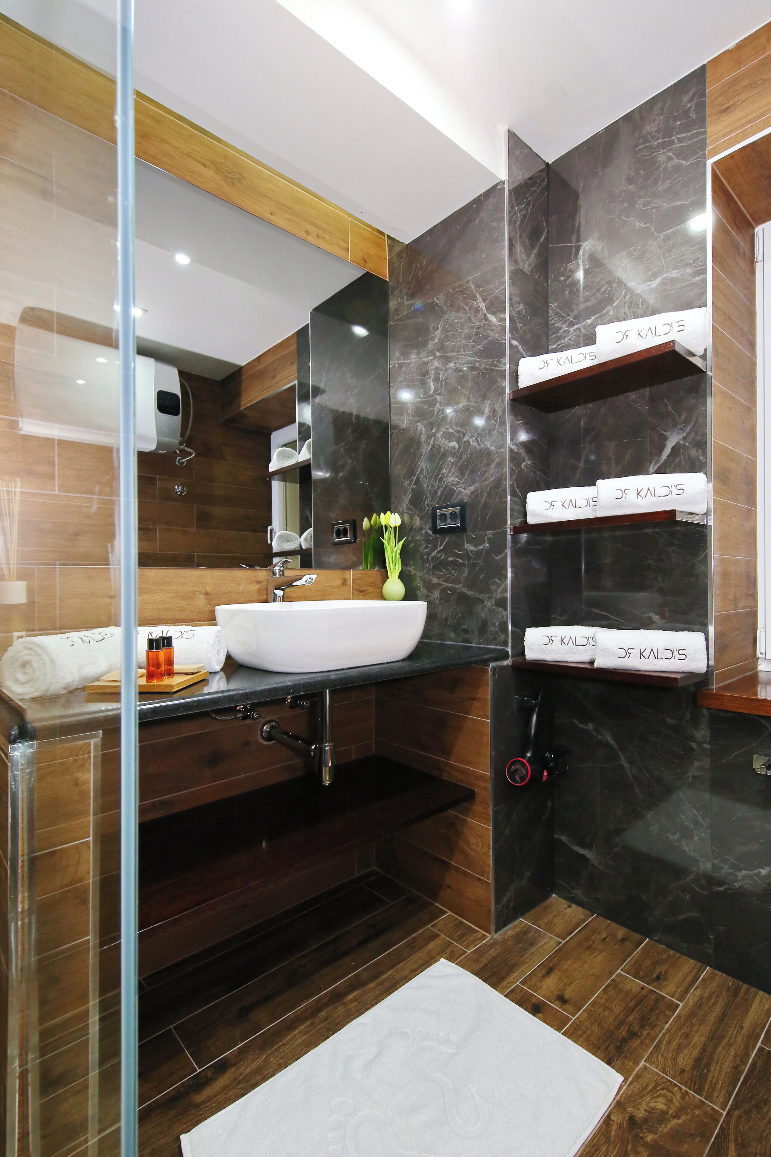A modern bathroom with a large mirror, a white sink, and dark marble walls. There are towels hanging on a shelf and a glass shower enclosure.