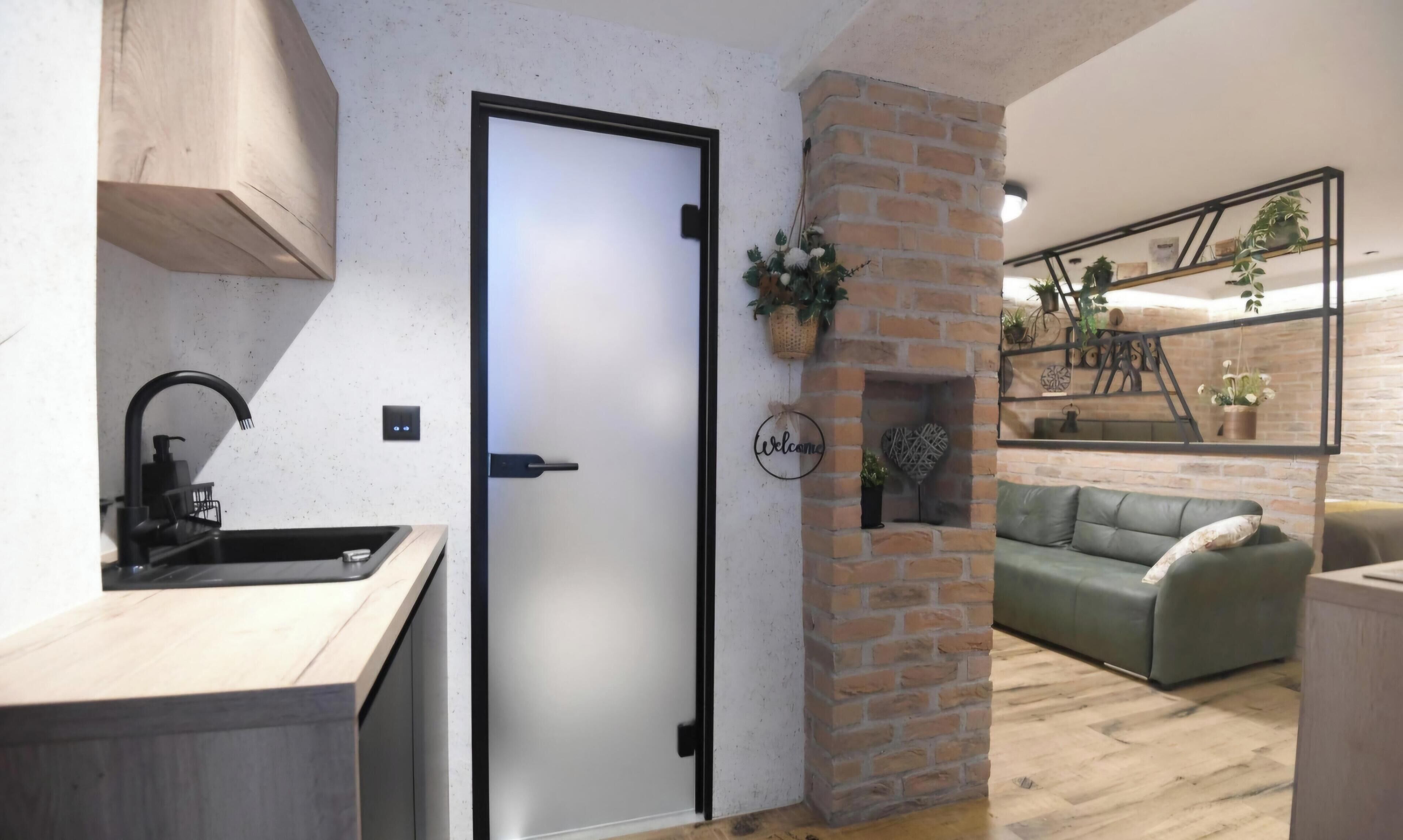 A modern kitchen with a built-in sink and a door leading to a living area with a green sofa and a brick accent wall.