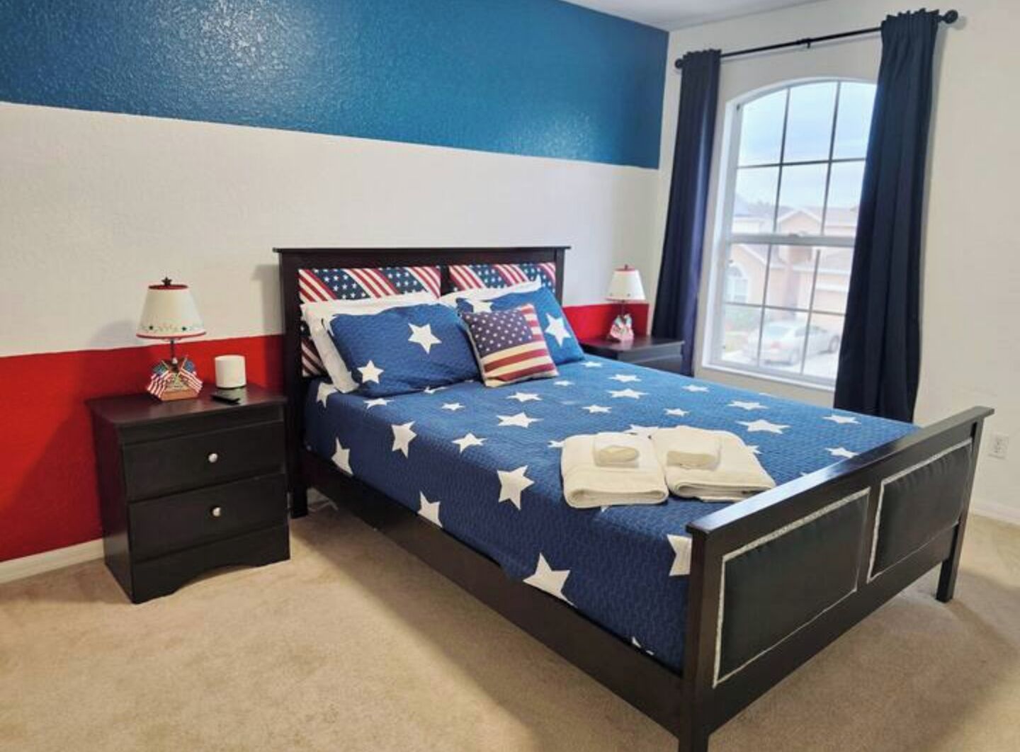 A bedroom with a bed featuring a star-patterned bedspread, a nightstand with a lamp, and a window with curtains.