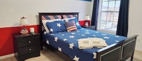 A bedroom with a bed featuring a star-patterned bedspread, a nightstand with a lamp, and a window with curtains.