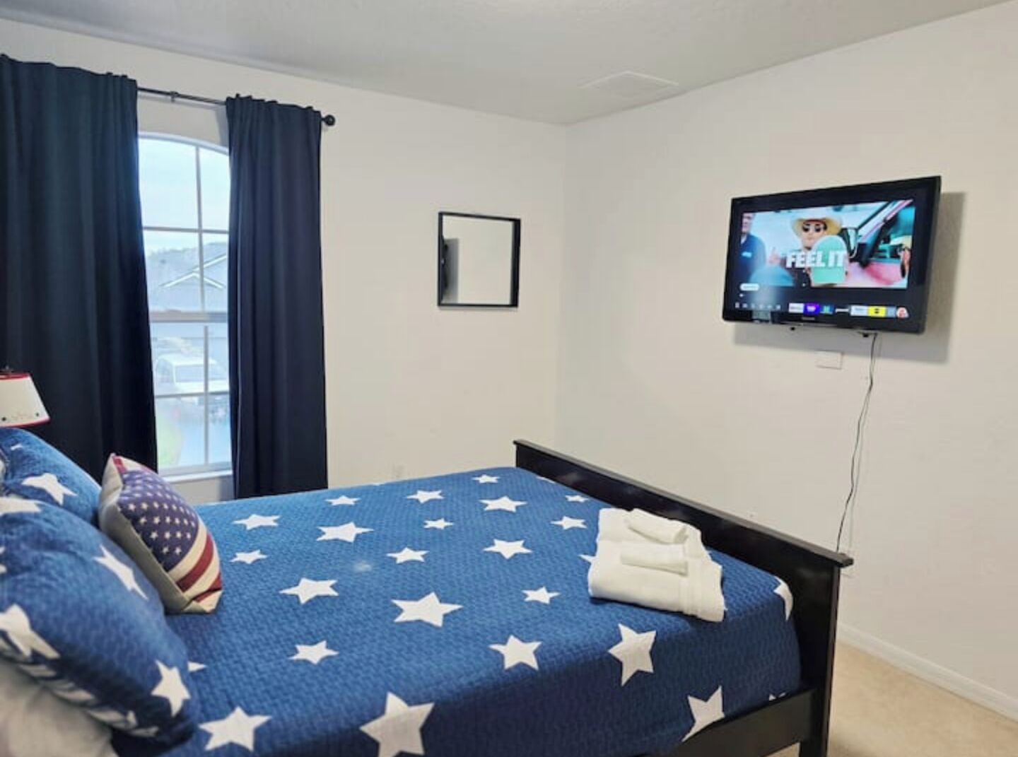 A hotel room with a bed featuring a blue blanket with white stars, a television mounted on the wall, and a window with dark curtains.