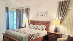Une chambre à coucher avec un lit en bois, deux tables de chevet avec des lampes, un fauteuil et une vue sur l’extérieur grâce à de grandes fenêtres.