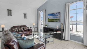 Un vaste salon comprenant un canapé en cuir marron, une table basse en verre et un téléviseur à écran plat fixé au mur. On y trouve deux tables d’appoint avec des lampes, ainsi qu’une grande fenêtre agrémentée de rideaux.