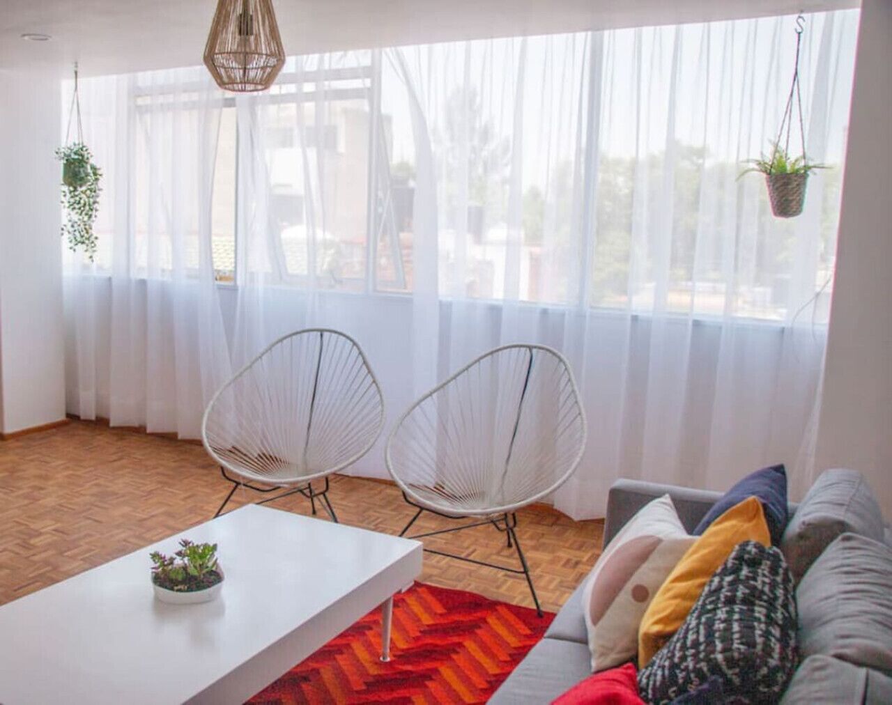 Un salón con un sofá blanco, dos sillas de estructura metálica blanca, una mesa de centro blanca y un tapete de diseño rojo y naranja.