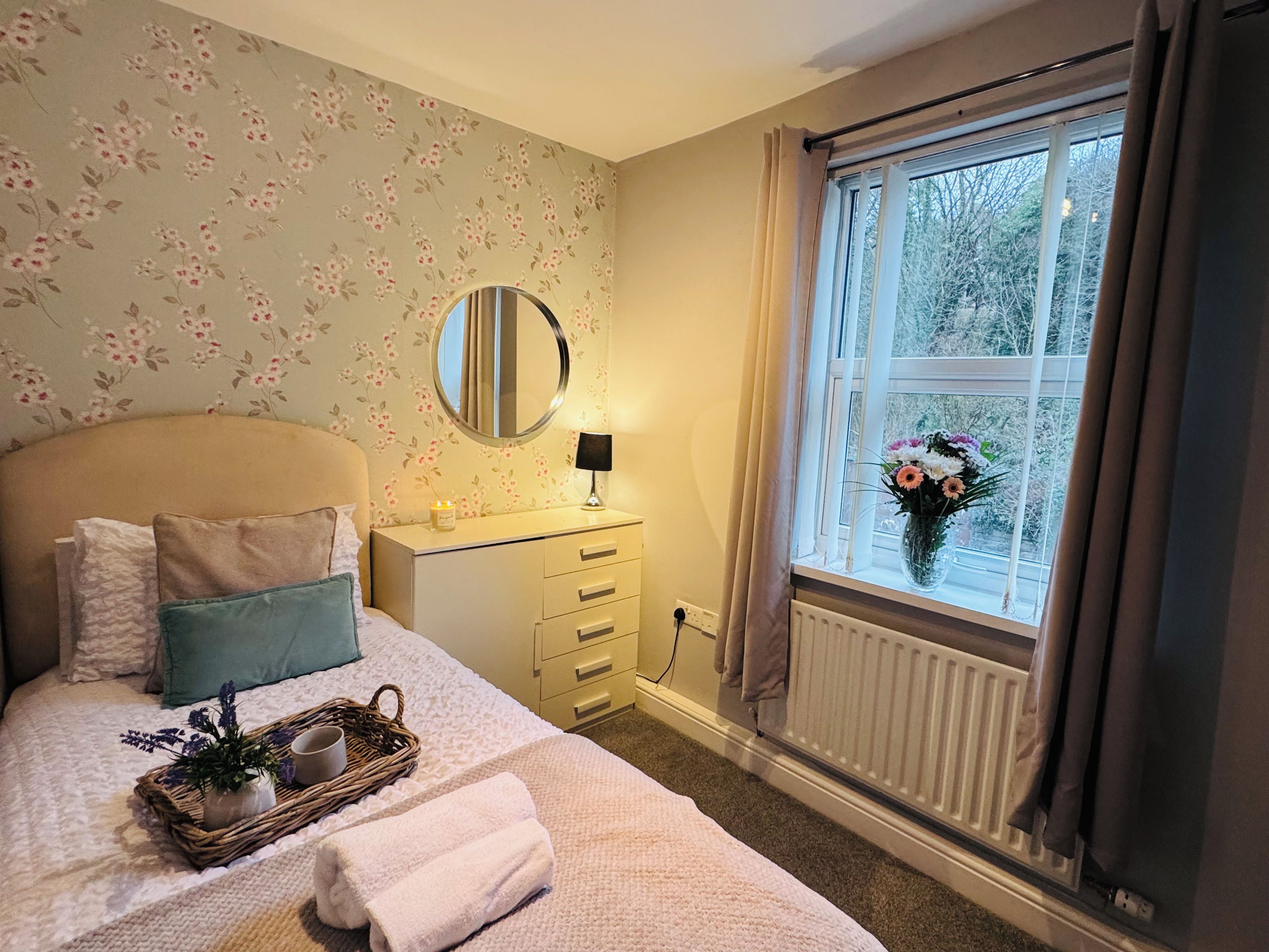 Un dormitorio con cama, mesita de noche con lámpara, espejo, ventana con cortinas y radiador.