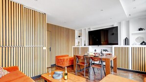 A modern living room with a wooden slat wall, a dining area with a wooden table and chairs, and a kitchen with a TV and white cabinets.