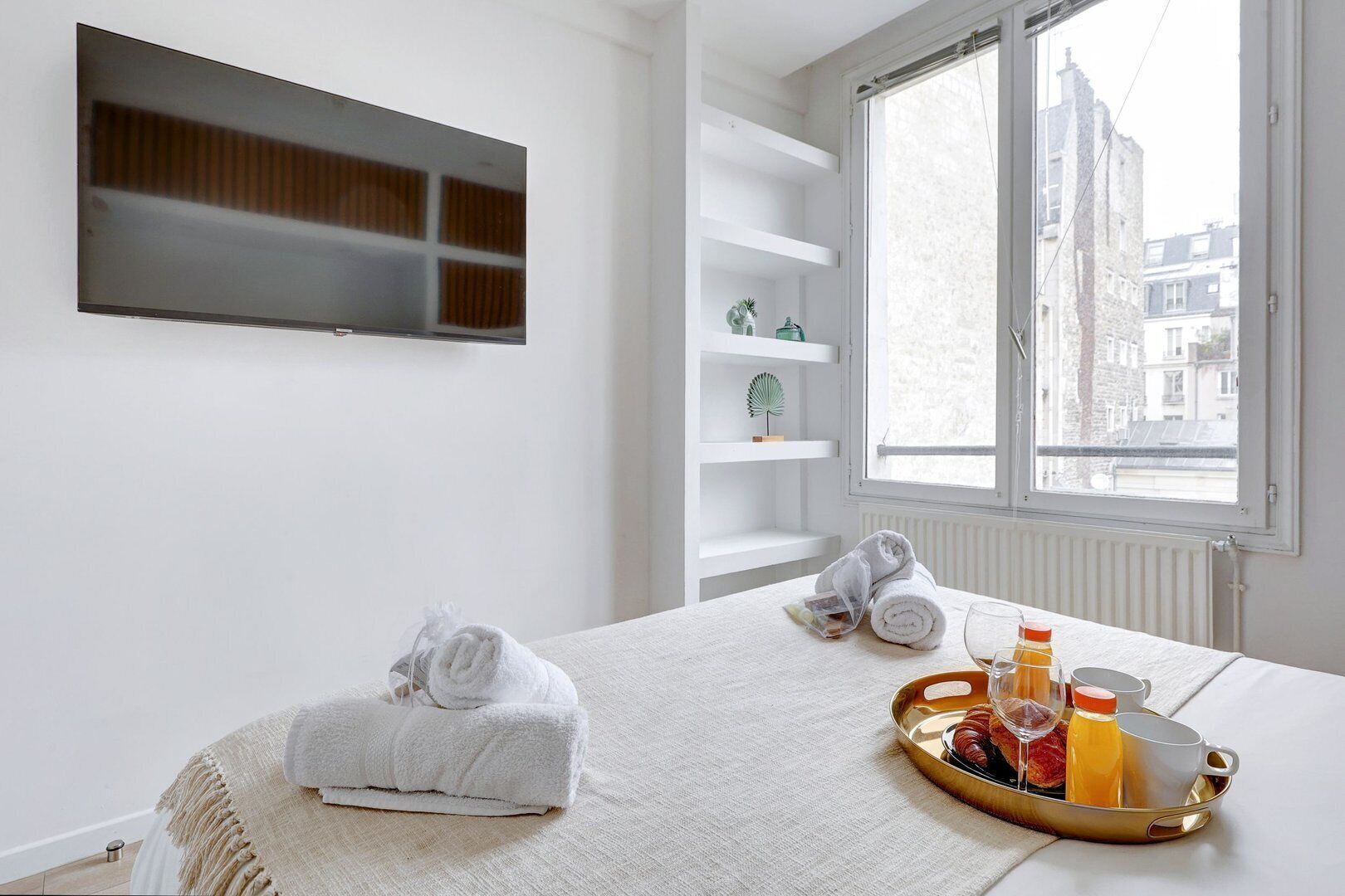 A hotel room with a bed, a window with a view of buildings, a shelf with decorative items, and a tray with drinks and a glass.