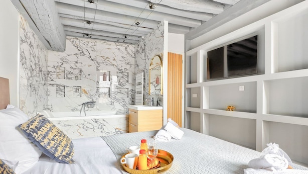 A modern hotel room with a marble-patterned wall, a large bed with a tray of breakfast items, a bathroom with a large mirror, and a view of the city.