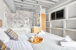 A modern hotel room with a marble-patterned wall, a large bed with a tray of breakfast items, a bathroom with a large mirror, and a view of the city.