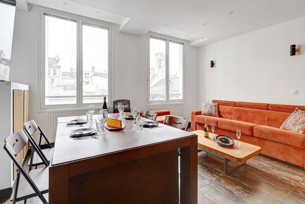 Una sala moderna con una mesa de comedor de madera dispuesta para cuatro personas, un sofá naranja vibrante y ventanas amplias con vistas a la ciudad.