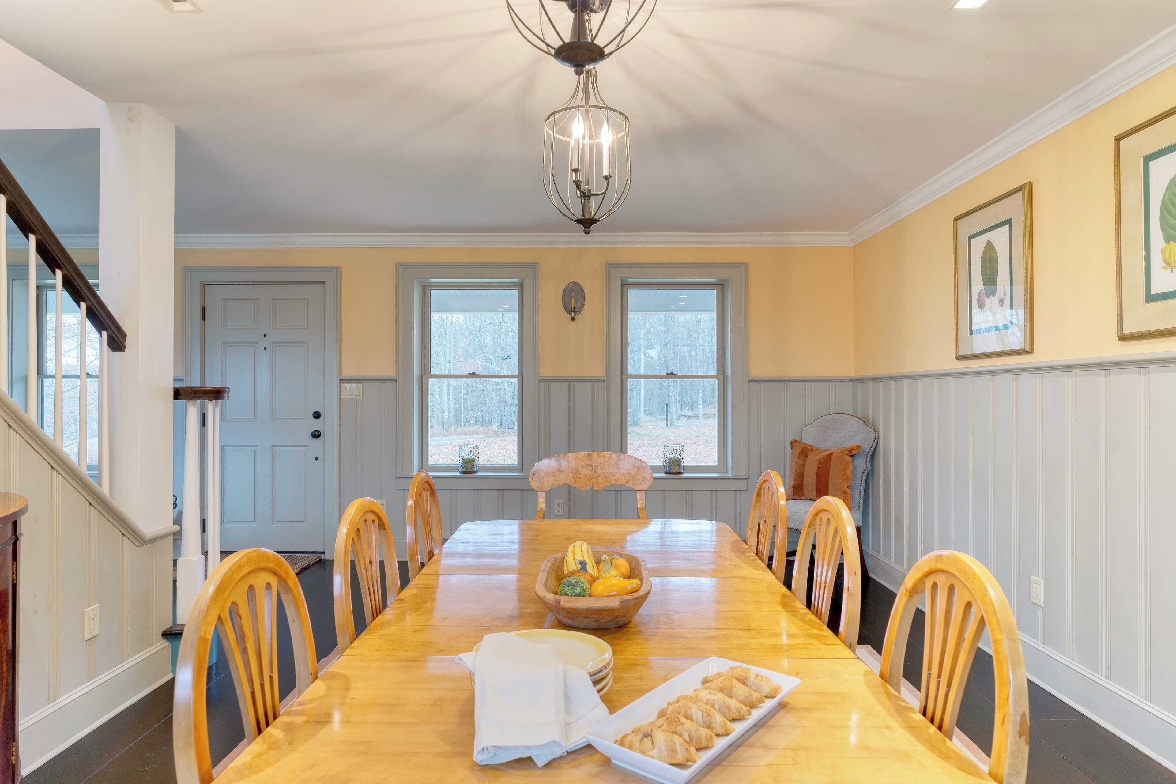 A dining room with a wooden table, chairs, a chandelier, and a window with a view of trees.