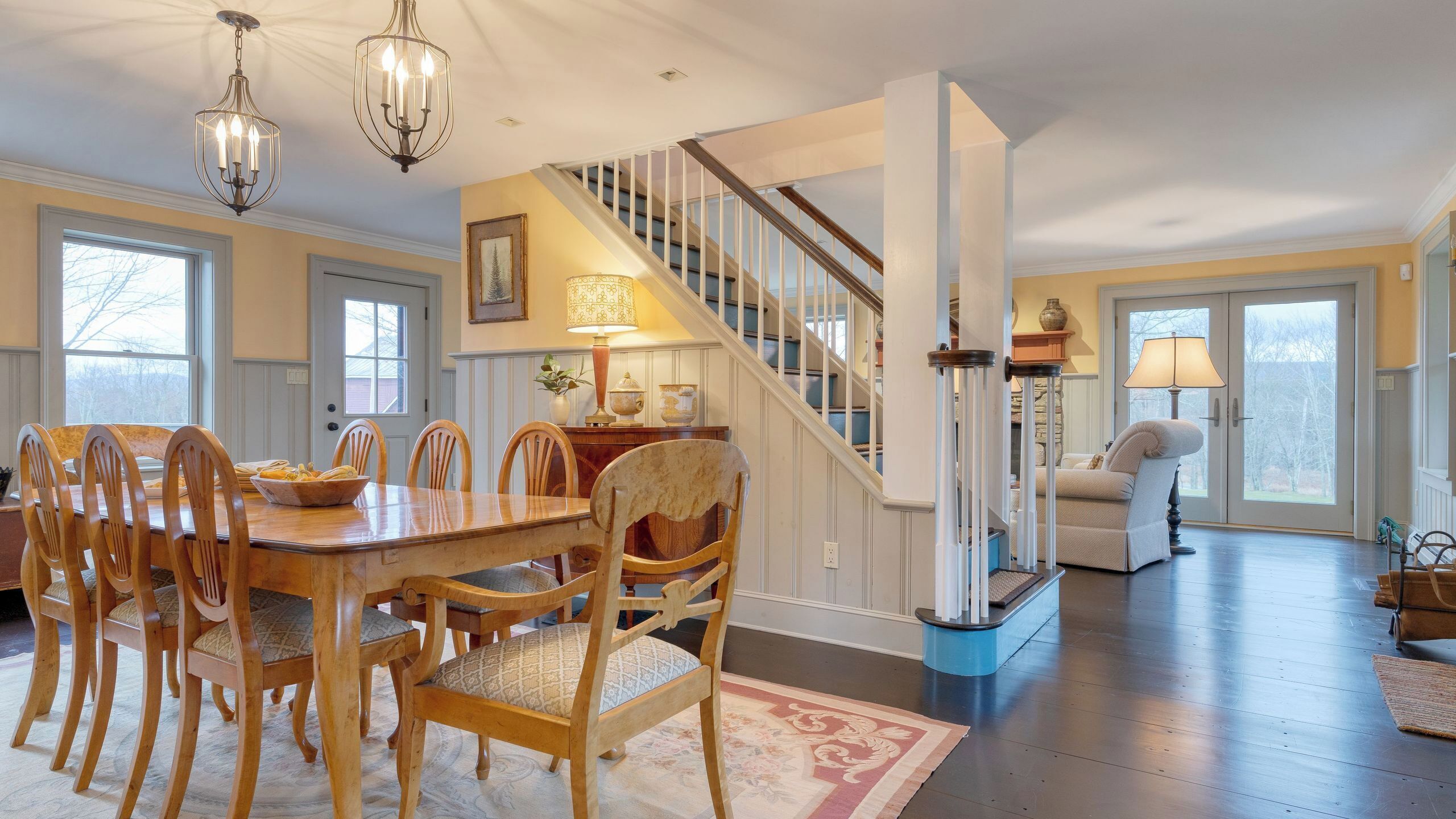 A dining area with a wooden table and chairs, a staircase with a railing, and a living room with a sofa and lamp.