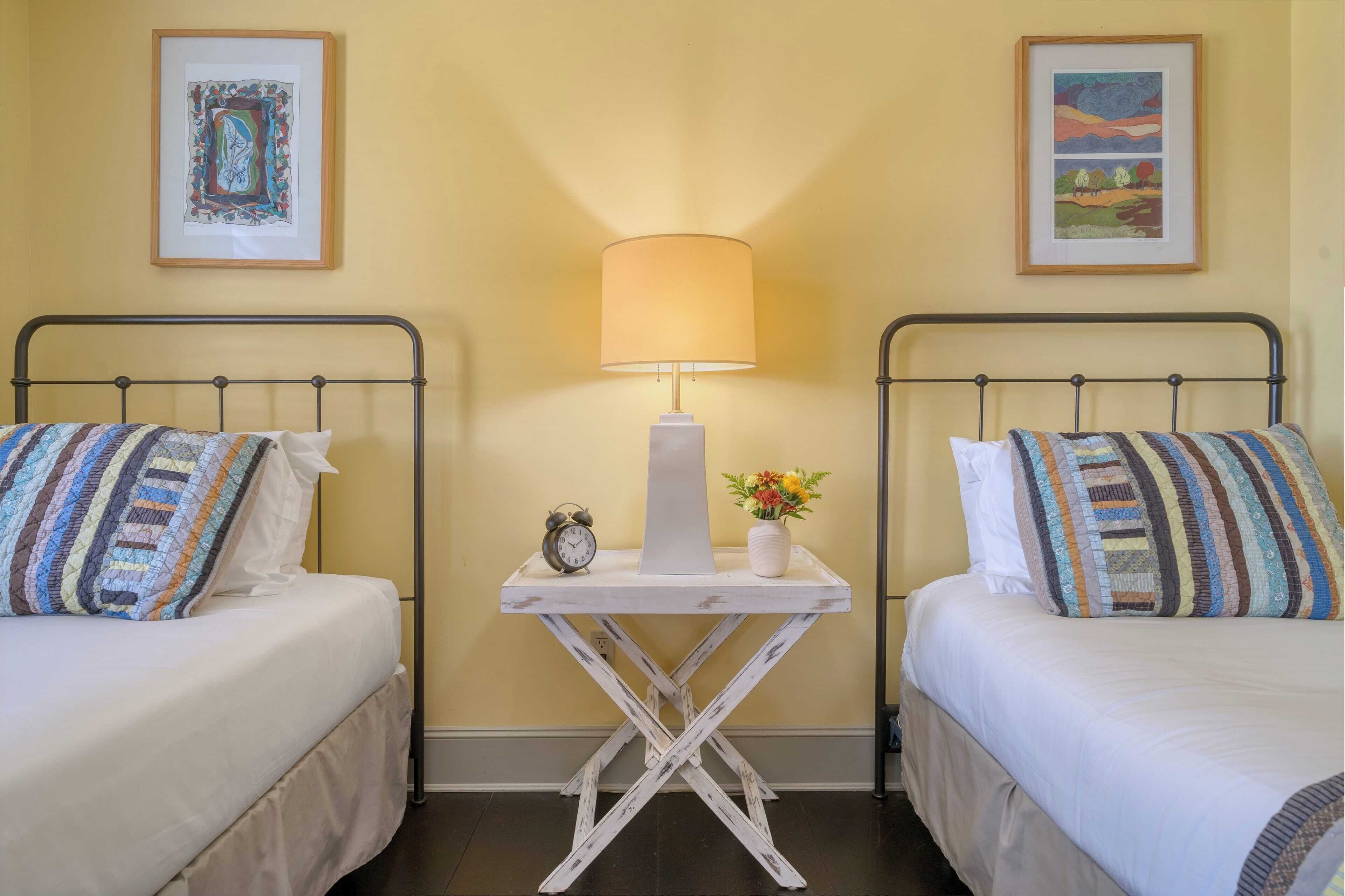 A hotel room with two beds, a bedside table with a lamp, a clock, and a vase with flowers.