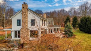 A two-story house with a stone chimney, a porch with outdoor seating, and a well-maintained lawn. - Catskill Park Farm by Avantstay Huge Yard, Fire Pit Near Windham Ski Resort (Ashland)