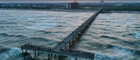 A pier extending into a body of water with waves, a distant building, and a sunset in the background.