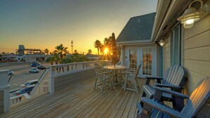 A wooden deck with outdoor seating, a view of the sunset, and a building with a sloped roof.