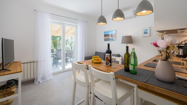 A modern kitchen with a dining table, chairs, and pendant lights. There is a TV on a shelf, a window with curtains, and a view of the outdoors. - Attractive Apartment With Garden (Porec)
