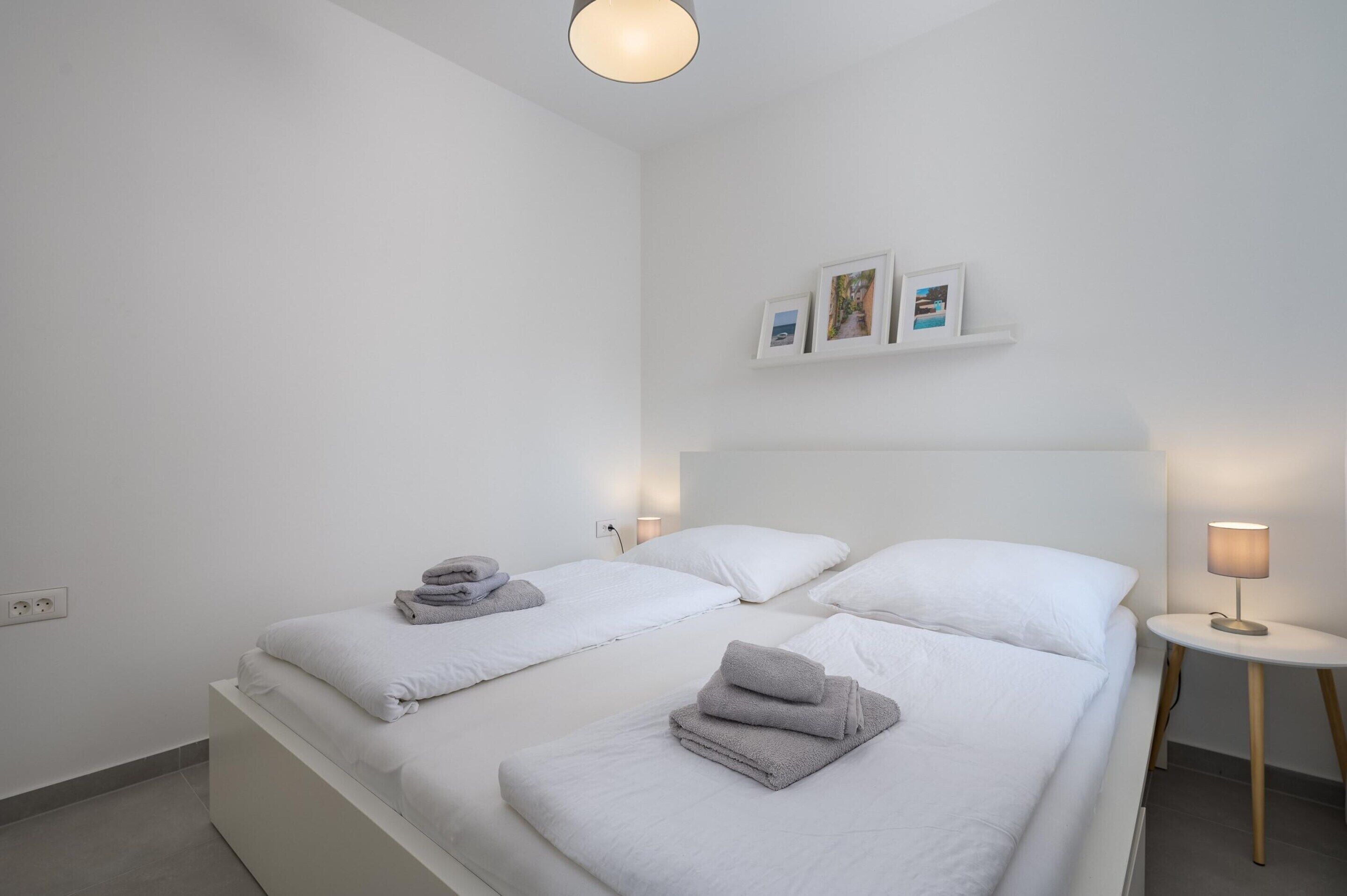 Habitación de hotel con dos camas, ropa de cama blanca, una mesita de noche con lámpara y una estantería en la pared con fotografías.