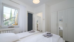A hotel room with two beds, a window with a view of a staircase and trees, a white wardrobe, and a bathroom with a sink and mirror. - Attractive Apartment With Garden (Porec)