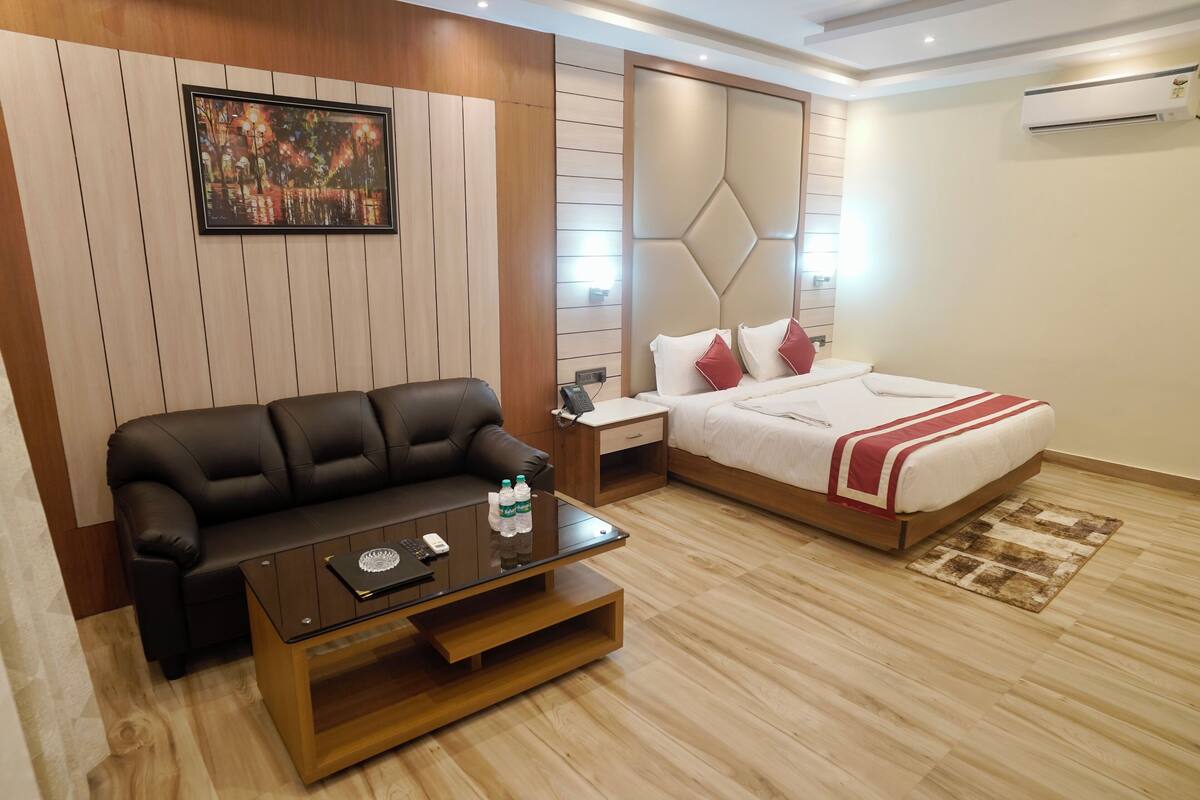 Habitación de hotel moderna con pared de paneles de madera, un sofá de cuero negro, una mesa de centro de vidrio, una cama con ropa de cama blanca y almohadas rojas, y una mesita de noche pequeña.