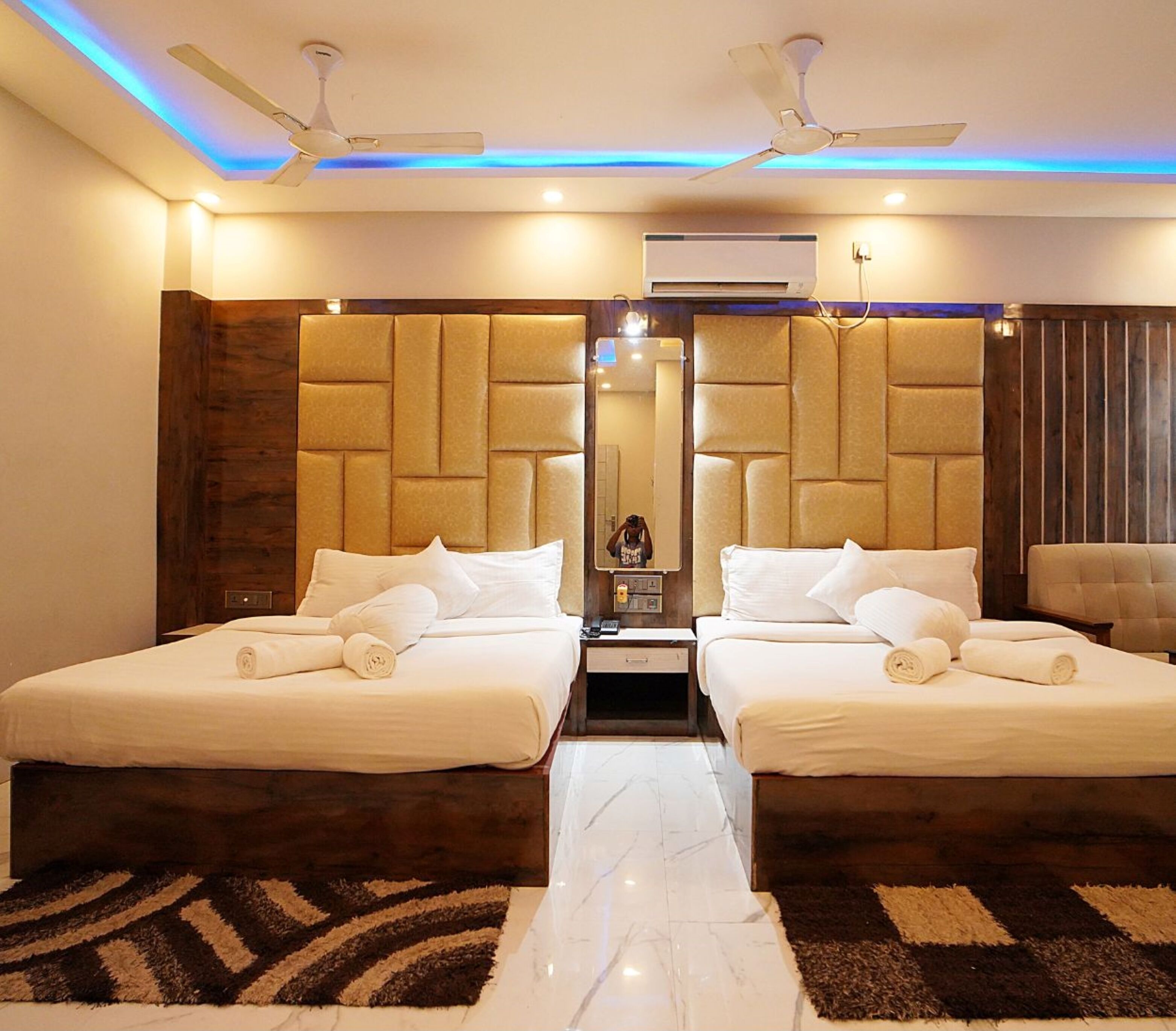 A hotel room with two beds, a central mirror, ceiling fans, and an air conditioning unit.