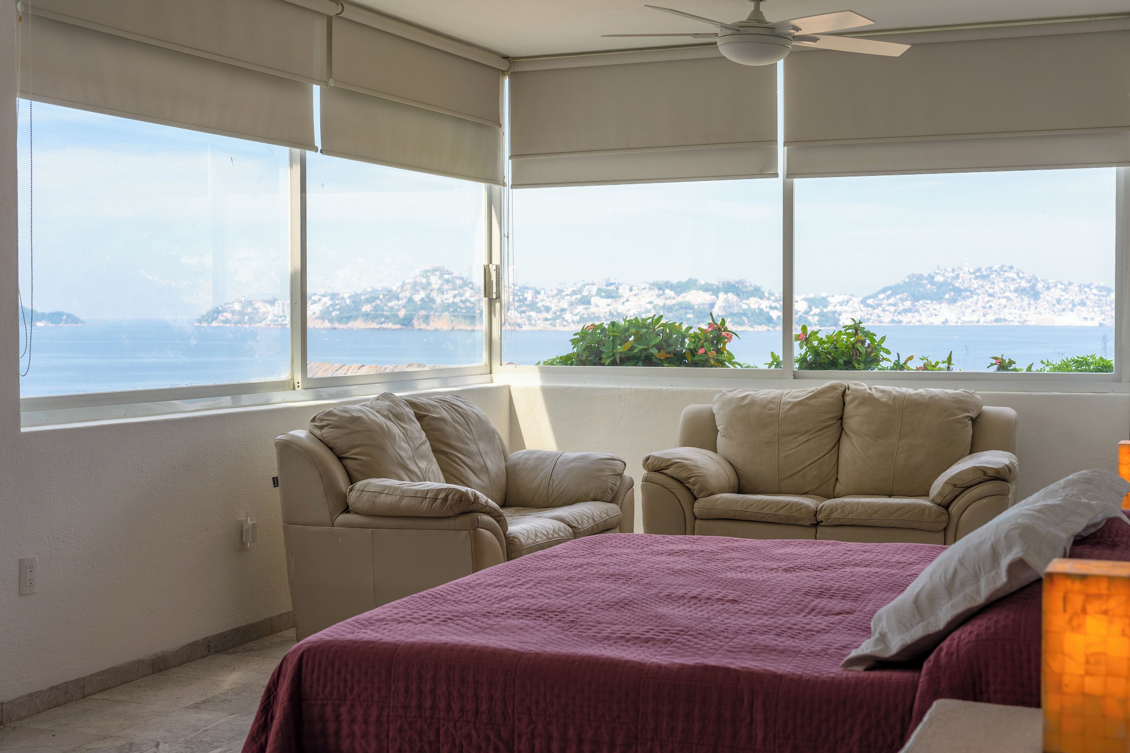 Un dormitorio con una cama, dos sillones color beige, un ventilador de techo y amplios ventanales que ofrecen vista al mar y a una colina.