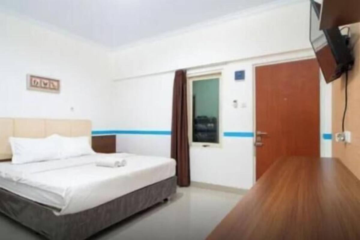 A hotel room with a bed, a wooden desk, a television mounted on the wall, and a door leading to another room.