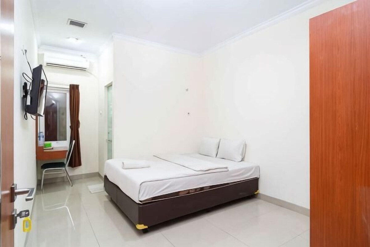 A single-bed room with a dark-colored bed frame, a desk with a chair, a television mounted on the wall, and an air conditioning unit.