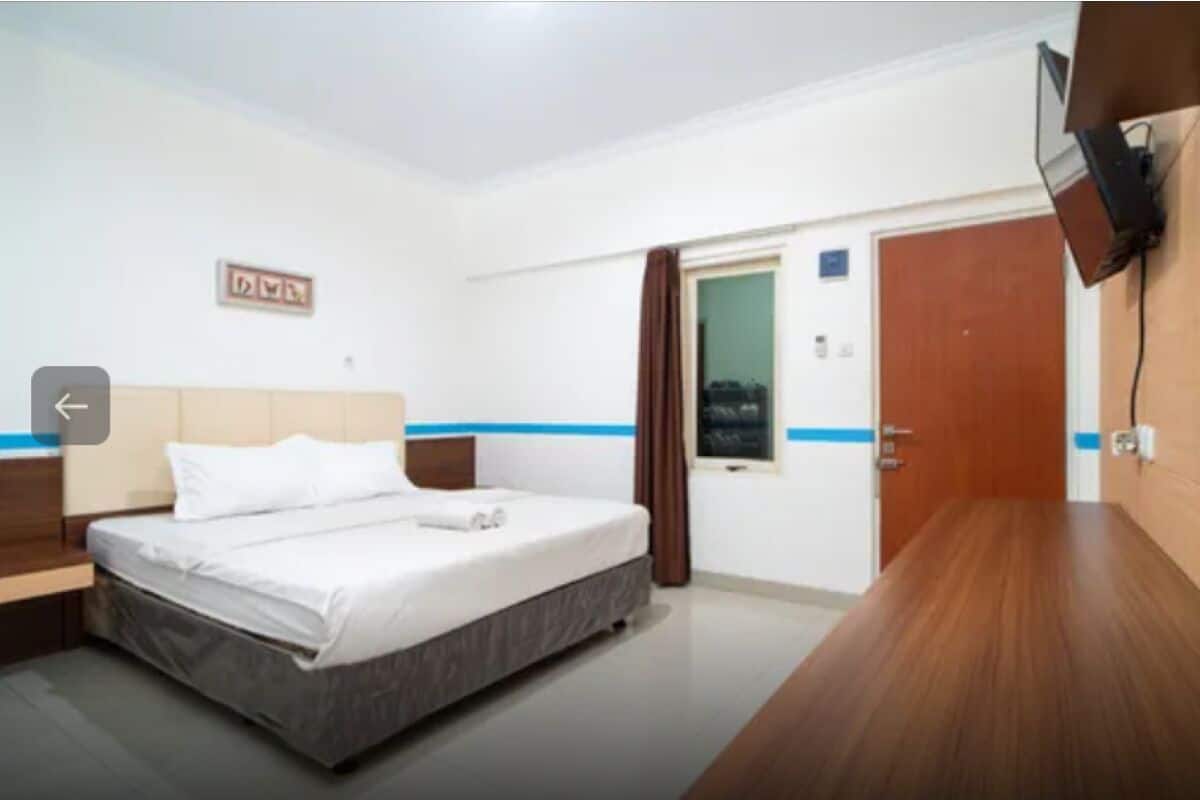 A hotel room with a bed, a wooden nightstand, a flat-screen TV mounted on the wall, and a door with a window.