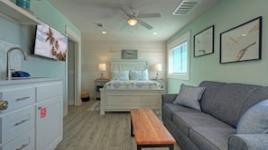 A bedroom with a bed, a gray sofa, a wooden bench, a ceiling fan, and a window with blinds. - Dolphin at Trout Street Queen Suite w/ Kitchenette Marina-front + Patio (Port Aransas)