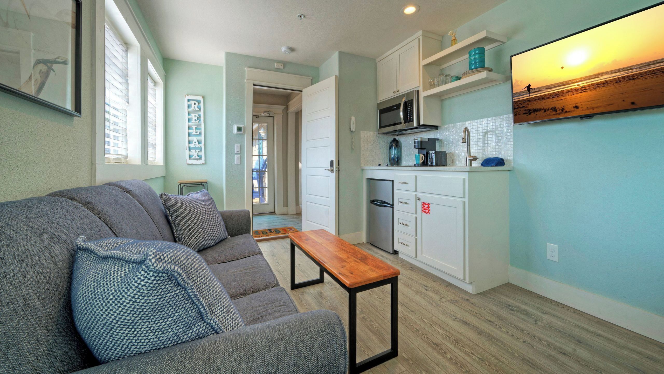A compact living space with a sofa, a small wooden table, a kitchenette, and a view of the beach.