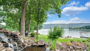 A serene lakeside setting with a small waterfall, lush greenery, and a view of the lake and distant hills. - Hook & Paddle - Luxury home on SML with kayaks and private dock. (Huddleston)