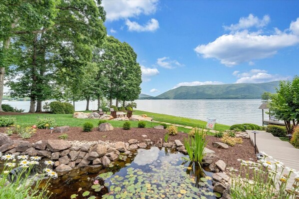 Un jardin au bord du lac, avec un étang, un espace détente et une terrasse en bois offrant une vue sur l’eau.