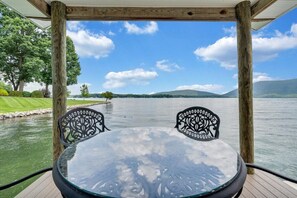 Un espace extérieur aménagé, abrité et doté d’une table et de deux chaises, offrant une vue sur un lac paisible et sur une chaîne de montagnes lointaine.